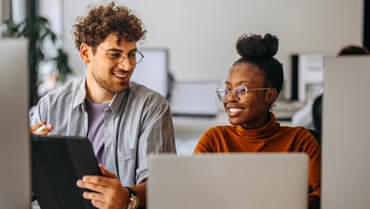 Zwei junge Berufspersonen, eine Frau und ein Mann, arbeiten zusammen an einem modernen Arbeitsplatz. Beide sitzen vor Computern und lachen, während sie auf ein digitales Gerät schauen. Sie diskutieren offensichtlich ein Thema im Bereich Marketing- und Vertriebsautomatisierung.