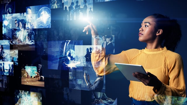 A woman in a yellow blouse interacts with a virtual display of various digital images and icons, possibly representing data, technology, and global connections. She holds a tablet and gestures towards the holographic images, surrounded by a futuristic interface. | AI for HR SUNZINET