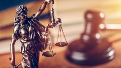 A Justitia statue with scales and blindfold in the foreground, symbolizing justice and the rule of law. In the background, a slightly blurred wooden gavel rests on a book. The image represents the Accessibility Strengthening Act (BFSG) 2025 and its legal implications.