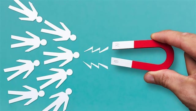 A hand holds a red and white horseshoe magnet aimed at several stylized white paper figures. Jagged lines illustrate the magnetic pull toward the figures. The background is solid turquoise. The image symbolizes customer retention