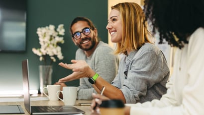 Symbolbild zu Interne Kommunikation Trends in 2025: Ein Team in einem modernen Büro bei einer Besprechung. Eine Frau spricht gestikulierend, während ihre Kollegen aufmerksam zuhören und lächeln. Laptops, Notizbücher und Kaffeetassen auf dem Tisch unterstreichen die produktive Atmosphäre.