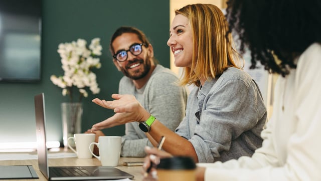 Symbolbild zu Interne Kommunikation Trends in 2025: Ein Team in einem modernen Büro bei einer Besprechung. Eine Frau spricht gestikulierend, während ihre Kollegen aufmerksam zuhören und lächeln. Laptops, Notizbücher und Kaffeetassen auf dem Tisch unterstreichen die produktive Atmosphäre.