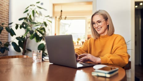 Das Bild zeigt eine junge Frau, die in einem modernen, gut beleuchteten Raum am Schreibtisch sitzt und auf ihrem Laptop arbeitet. Sie trägt ein orangefarbenes Sweatshirt und lächelt, während sie mit dem Laptop interagiert. Im Hintergrund sind Pflanzen und ein Fenster zu sehen. Die Frau könnte an der Planung oder Umsetzung von B2B Marketing Automation arbeiten, um Marketingprozesse in Unternehmen zu optimieren und zu automatisieren.