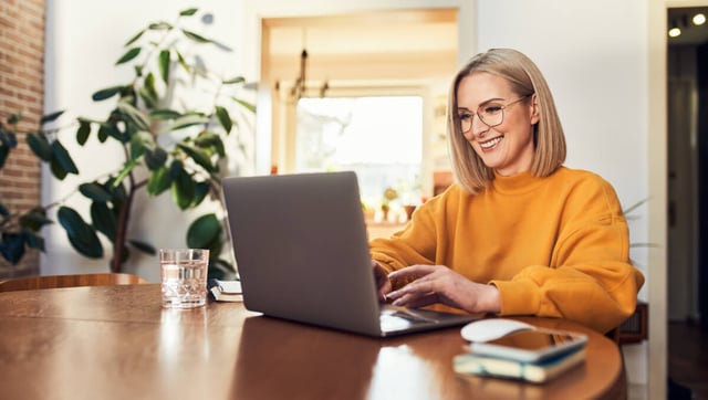 Das Bild zeigt eine junge Frau, die in einem modernen, gut beleuchteten Raum am Schreibtisch sitzt und auf ihrem Laptop arbeitet. Sie trägt ein orangefarbenes Sweatshirt und lächelt, während sie mit dem Laptop interagiert. Im Hintergrund sind Pflanzen und ein Fenster zu sehen. Die Frau könnte an der Planung oder Umsetzung von B2B Marketing Automation arbeiten, um Marketingprozesse in Unternehmen zu optimieren und zu automatisieren.
