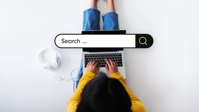 A person wearing a yellow sweater is sitting on the floor with a laptop on their lap. Their hands are typing on the keyboard. Overlaid is a graphical search bar with the word 'Search ...' symbolizing onsite search. Next to it is a green magnifying glass icon. To the left of the person, white headphones are placed on the floor.